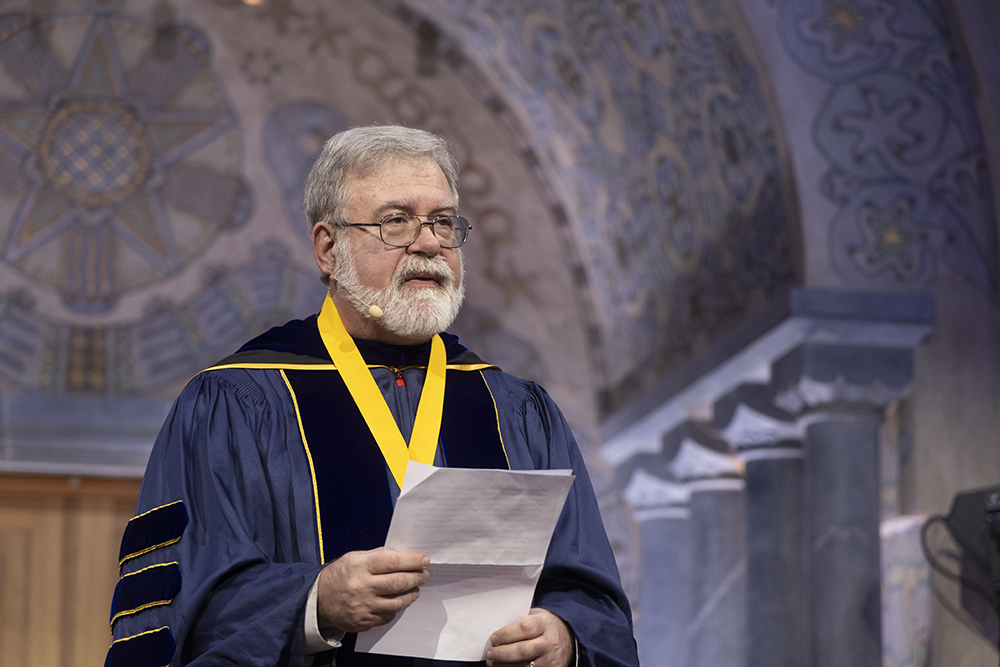Old man in glass and graduation robes holds a paper and gives a speech.